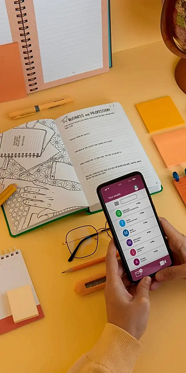 Hands holding a phone testing an application with an open notebook on a yellow desk.